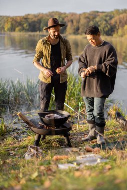 Two multiracial friends cooking Ukha soup in cauldron on bonfire during a picnic. Men talking and having fun together on lake coast. Leisure, weekend and vacation in nature