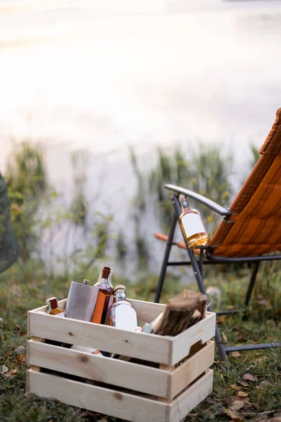 Bottles with strong alcohol at wooden box and folding chair at picnic or fishing. Bottle with blank label to copy paste. Mockup image for branding drink