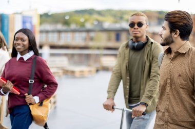 Gülümseyen çok ırklı öğrenciler üniversite kampüsünde birlikte yürürler. Eğitim ve öğrenim kavramı. Öğrenci yaşam tarzı ve arkadaşlık. Siyah adam elektrikli scooter kullanıyor. Modern ulaşım fikri