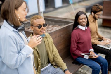 Çok ırklı öğrenciler dışarıdaki ahşap bankta otururken dizüstü bilgisayardan bir şey izliyorlar. Uzaktan kumanda ve e-öğrenme kavramı. Öğrenci yaşam tarzı fikri. Üniversite kampüsündeki genç kızlar ve erkekler