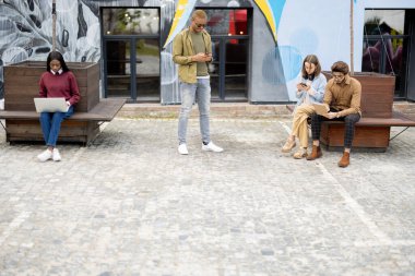 Multiracial students with digital gadgets outdoors. Concept of students lifestyle. Idea of remote and e-learning. Young guys and girls at university campus