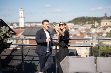 Beyaz kadın ve Asyalı adam balkonda akıllı telefondan selfie çekiyorlar. Gülümseyen çift birlikte eğleniyor. Güneşli bir gün. İnsanların arka planında şehir görünümü