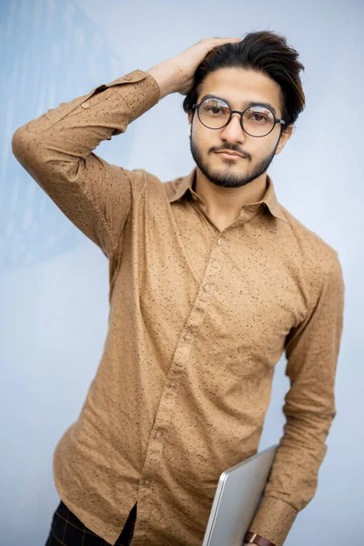 Young indian man with laptop looking at camera. Handsome smiling stylish guy in glasses wearing t-shirt. Male person on blue background