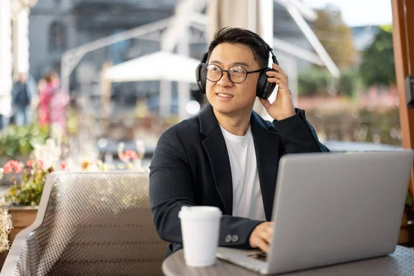 Asian businessman in earphones typing on laptop during work in cafe. Concept of remote and freelance work. Focused adult successful man wearing suit and glasses sitting at table with coffee