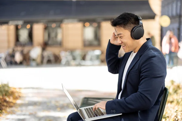 Irritated asian businessman enjoy music in earphones and working on laptop outdoors. Concept of remote work and freelance. Modern adult successful man wearing suit sitting on chair. Sunny day