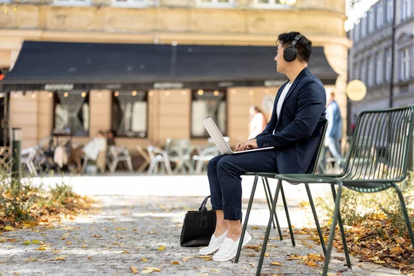 Irritated asian businessman in earphones working on laptop outdoors. Concept of remote work and freelance. Modern adult successful man wearing suit sitting on chair. Sunny day