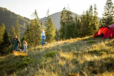 Young family with little girl walk together on green meadow while traveling with tent in the mountains during sunset. Happy family spending summer vacation at campsite