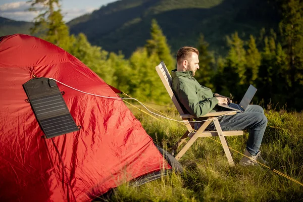 Man works on laptop while traveling with tent in the mountains. Charging computer with portable solar panel hanging on tent. Concept of remote work at campsite and renewable energy