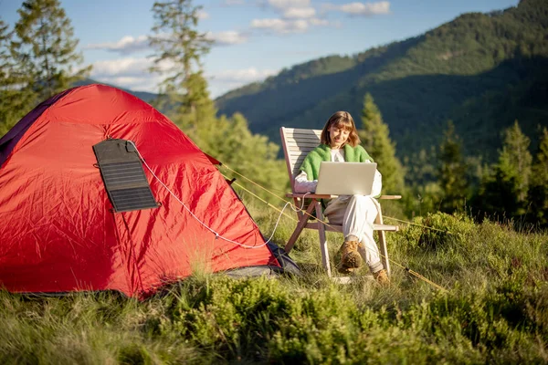 Young woman works on laptop while traveling with tent in the mountains. Charging computer with portable solar panel hanging on tent. Concept of remote work at campsite and renewable energy