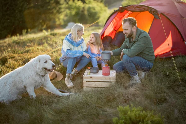 Couple with little girl and dog have picnic at campsite while traveling with tent in the mountains. Young family happily spendingvacations on nature