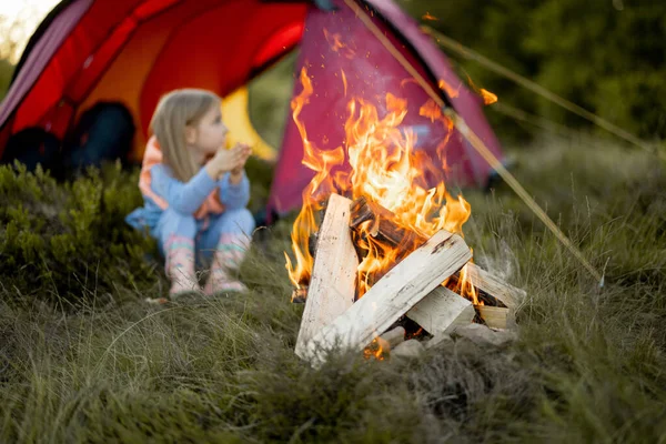 Little cute girl sit alone and enjoy campfire near tent on nature. Happy childhood and bonding with nature concept