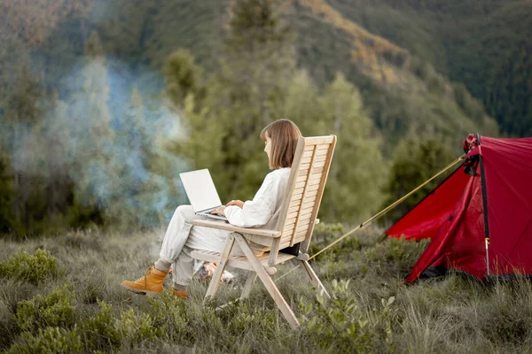 Young woman works on laptop while sitting relaxed on chair by the campfire, traveling with tent in the mountains. Concept of remote work and escape to nature