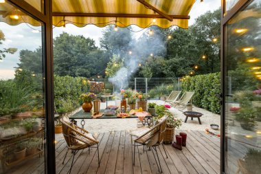 Atmospheric and cozy garden with dining place on terrace at dusk. Cooking food on disposable grill on beautiful table decorated with flowers. Wide view from the house