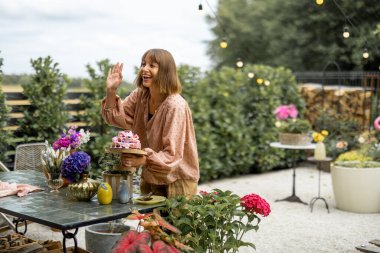Happy woman waving hand while putting festive cake on table decorated with flowers, preparing for the holiday in garden