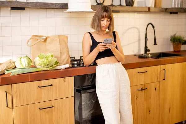 Young woman uses smart phone while standing with freshly bought groceries in kitchen at home. Concept of buying food online and mobile technologies