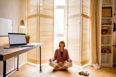 Young woman meditates after work, sitting on the floor at cozy home office. Stylish interior with workplace and computers in front. Mental health and relaxation, working from home concept