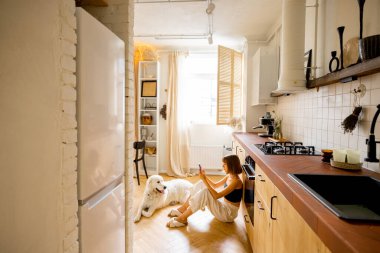 Woman uses smart phone while sitting with her dog in kitchen. Interior view on modern and stylish kitchen in beige tones. Domestic lifestyle concept