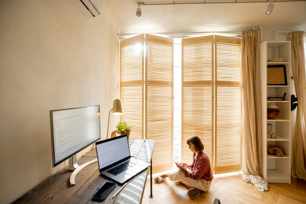 Woman sits on the floor by the window with a digital tablet, resting after work on computers at cozy and sunny home office. Wide interior view with workplace in front