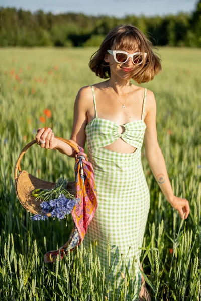 Stylish woman in dress walks with wild flowers on field, spending summertime carefree on nature