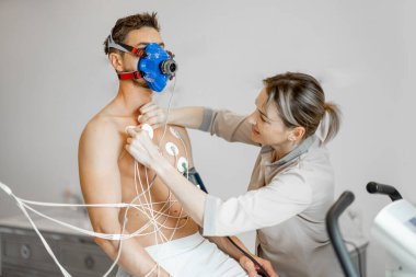 Nurse attaches electrodes to a man for a cardio endurance test during physical exercise on bike simulator, examining heart and vascular system. Man in breath mask on face