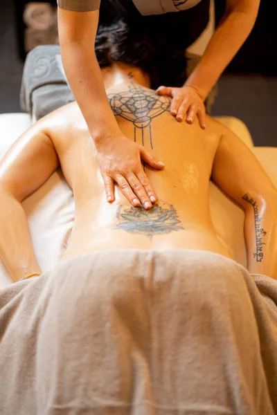 Woman receiving professional relaxing back massage at Spa salon. Client lying covered with towel on medical couch with tattoos on her back