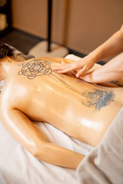 Woman receiving professional relaxing back massage at Spa salon. Client lying covered with towel on medical couch with tattoos on her back