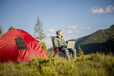 Adam dağlarda çadırla seyahat ederken dizüstü bilgisayarda çalışıyor. Çadırda asılı taşınabilir güneş paneli olan bir bilgisayar yükleniyor. Kamp alanında uzaktan çalışma ve yenilenebilir enerji kavramı