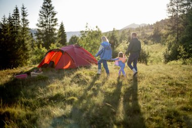 Genç beyaz bir aile ve küçük bir kız gün batımında dağlarda çadırla dolaşırken yeşil çayırlarda birlikte yürüyorlar. Mutlu aile yaz tatilini kamp alanında geçiriyor.