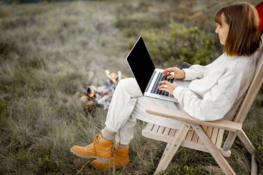 Young woman works on laptop while sitting relaxed on chair by the campfire, traveling with tent on nature. Concept of remote work