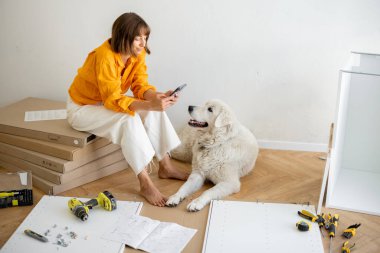 Young woman sits with her cute dog, using phone during repairing at new apartment. Assembling furniture by herself. DIY concept