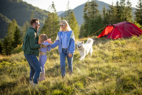 小さな女の子と犬と若い白人のカップルは 山の中でテントと一緒に旅行しながら楽しい時間を過ごしています キャンプ場で幸せな家族の夏休み 父は娘を腕に投げ — ストック写真