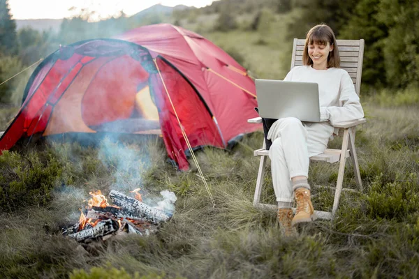 年轻女人坐在笔记本电脑上工作 坐在篝火边的椅子上放松 带着帐篷在山上旅行 远程工作和逃避现实的概念 — 图库照片