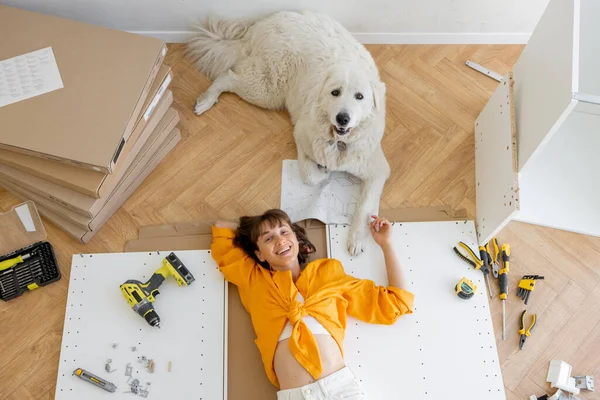 Young happy woman lies on floor with her cute dog, resting while making repairing at new apartment, top view. Young woman assembling furniture by herself. DIY concept