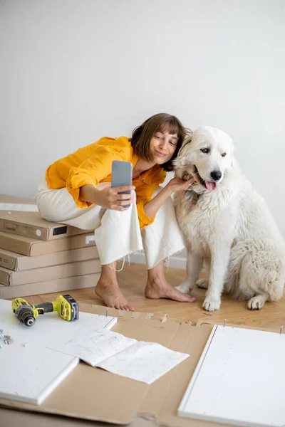 Young woman makes selfie photo on phone with her cute dog, have fun while making repairing at new apartment. Happy improvement and house repairing concept