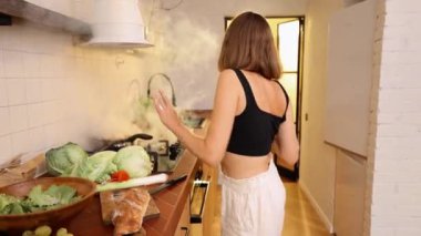 Kadın arka planda dumanla yanan tavaya rağmen mutfakta mutlu bir şekilde yemek pişiriyor. Sağlıklı vejetaryen yemekleri hazırlıyorum.