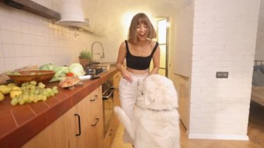 Kadın eğleniyor, arka planda yanmış yemeğin dumanıyla mutfakta yemek pişirirken köpeğiyle oynuyor. Genç ev hanımı ev işleri sırasında mutlu vakit geçiriyor.