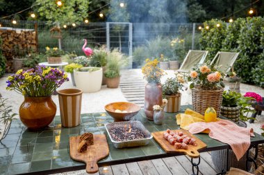 Atmospheric and cozy garden with dining place on terrace at dusk. Cooking food on disposable grill on beautiful table decorated with flowers