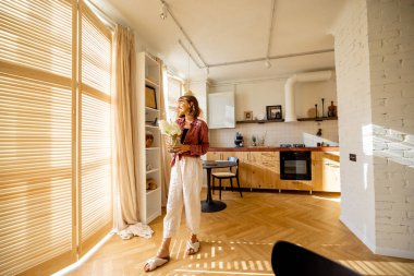 Woman walks with a vase of flowers, decoration and having some house chores at home. Wide interior view on sunny studio apartment in beige tones