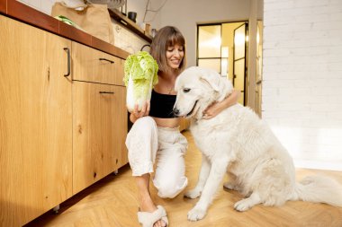 Young woman spends leisure time with her white dog while cooking healthy food in kitchen at modern apartment. Healthy eating and friendship with pets concept