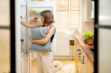 Young sleepy woman with a pillow takes some food from a fridge waking up at home. Night snacking and unhealthy lifestyle concept