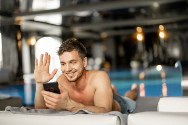 Man rests by the swimming pool indoors, talking on phone while lying relaxed on lounge bed indoors. Man spends holidays at spa resort with an indoor pool