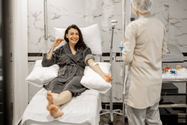 Woman in bathrobe sitting relaxed during blood washing procedure. Nurse preparing dropper and needle for the procedure at luxury medical centre