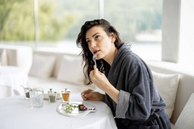 Beautiful woman in bathrobe enjoys delicious meal, sitting in white restaurant of haught cuisine during vacations at luxury spa resort