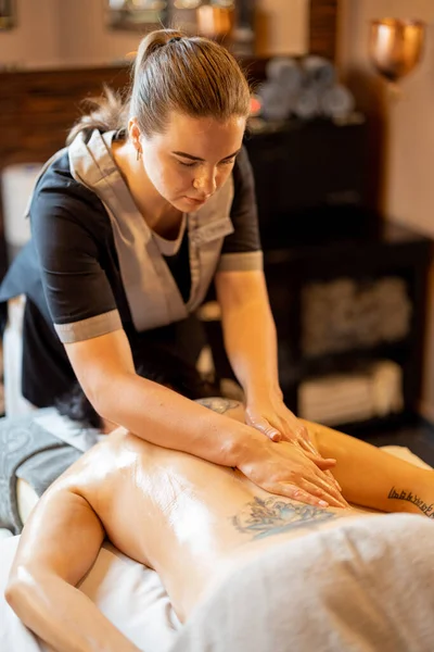 专业的女按摩师在温泉沙龙的按摩室为背部有纹身的妇女提供放松按摩 — 图库照片