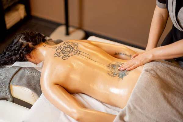 Woman receiving professional relaxing back massage at Spa salon. Client lying covered with towel on medical couch with tattoos on her back