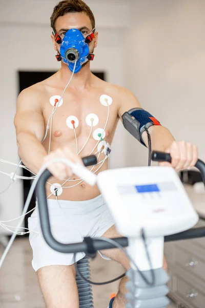 Man athlete with breath mask and electrodes training on bike simulator, examining his cardiovascular system at medical office