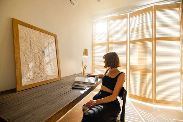 Woman reads some magazine while sitting at cozy workplace by the wooden table at home. Living room in beige tones with window blinds on background