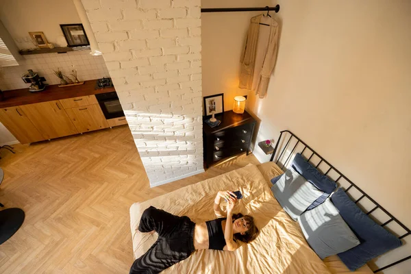 Vrouw Liggend Ontspannen Met Telefoon Bed Zonnige Studio Appartement Uitzicht — Stockfoto