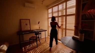 Kadın pencere perdelerini açar ve masanın yanına oturur. Modern ve şık dairesinde boş vakitlerini geçirir.