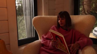 Kadın, doğadaki ahşap evde rahat bir kanepede otururken dergi okuyor. Camda, dağların manzaralı kamera hareketi